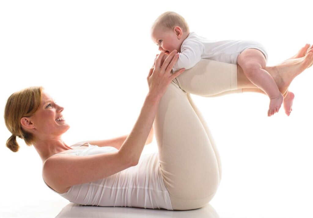 Young mother makes with her 6 month old daughter postnatal exercises, isolated against white background. Mama is down and has her baby lying on their lower legs.